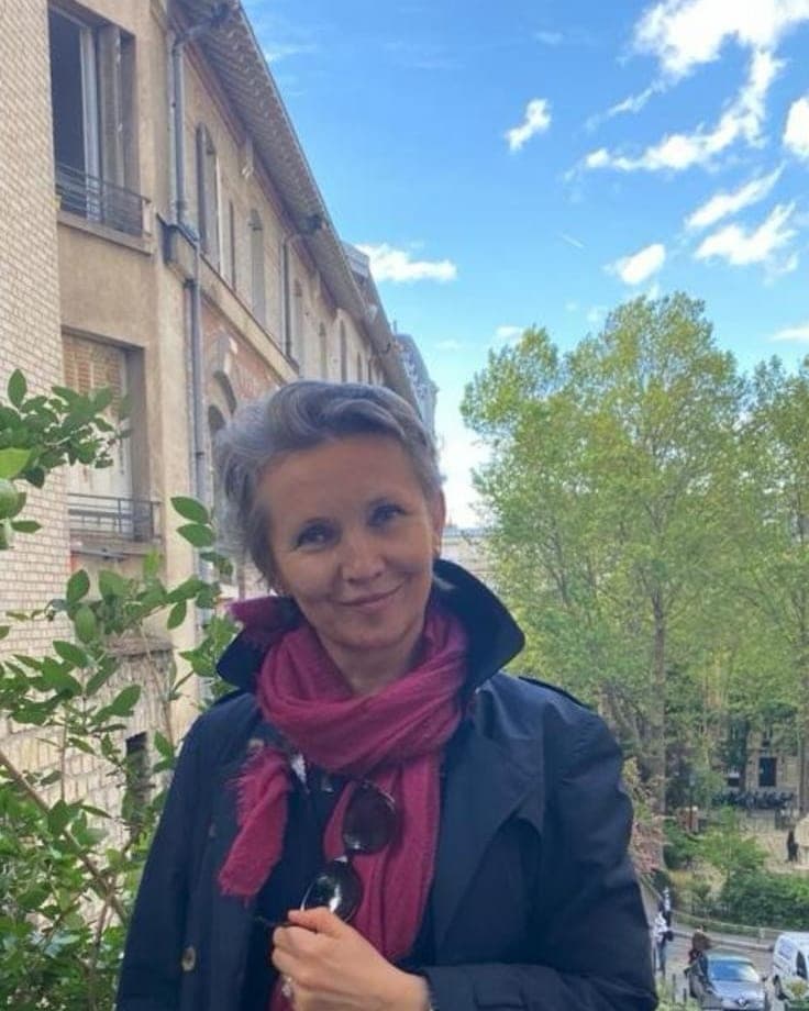 Portrait de Nataliya Yefimova en extérieur, manteau bleu et écharpe fuchsia, devant un immeuble et des arbres.